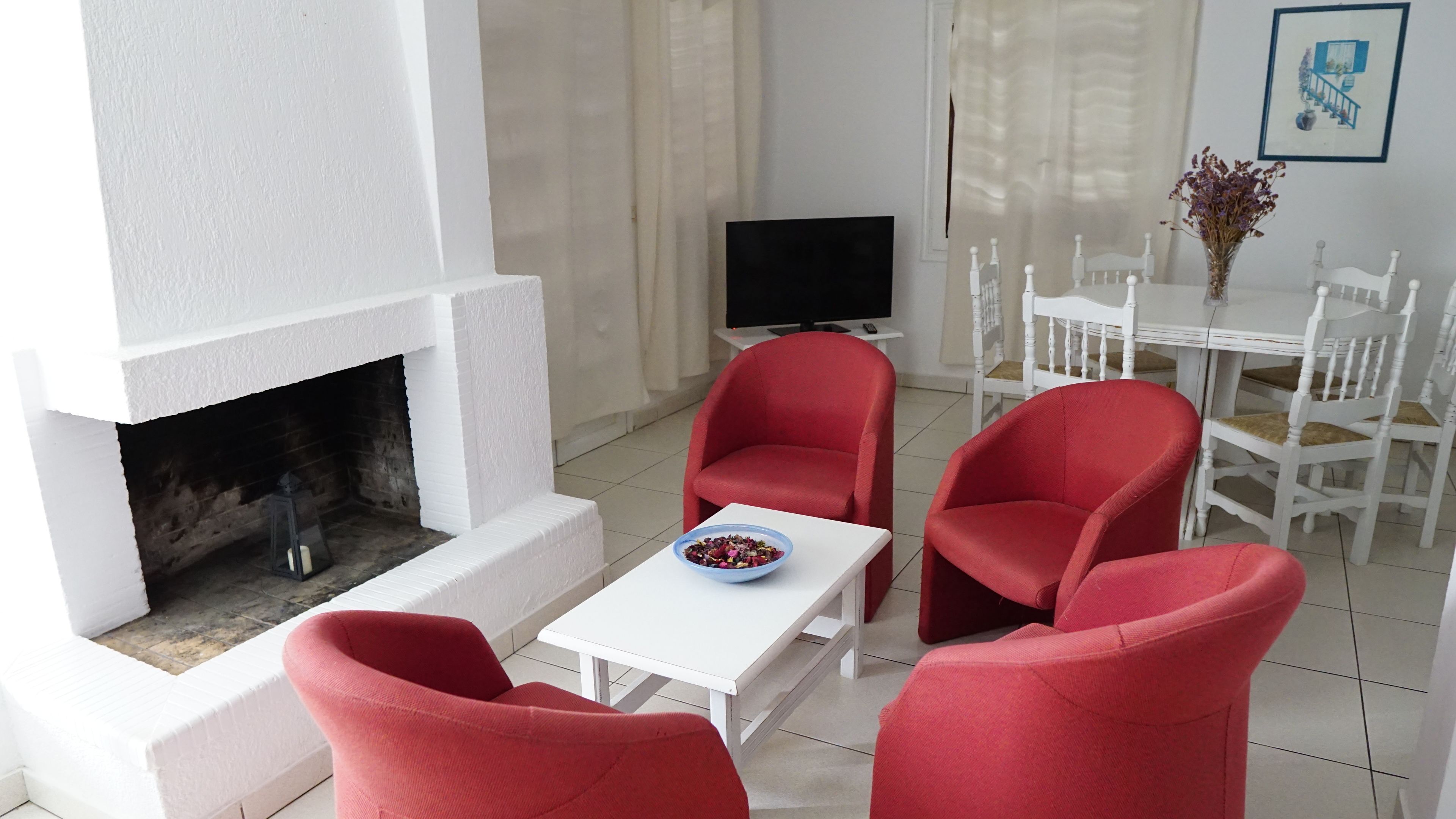 Un salon moderne avec une cheminée, des fauteuils rouges, une table blanche et un téléviseur.