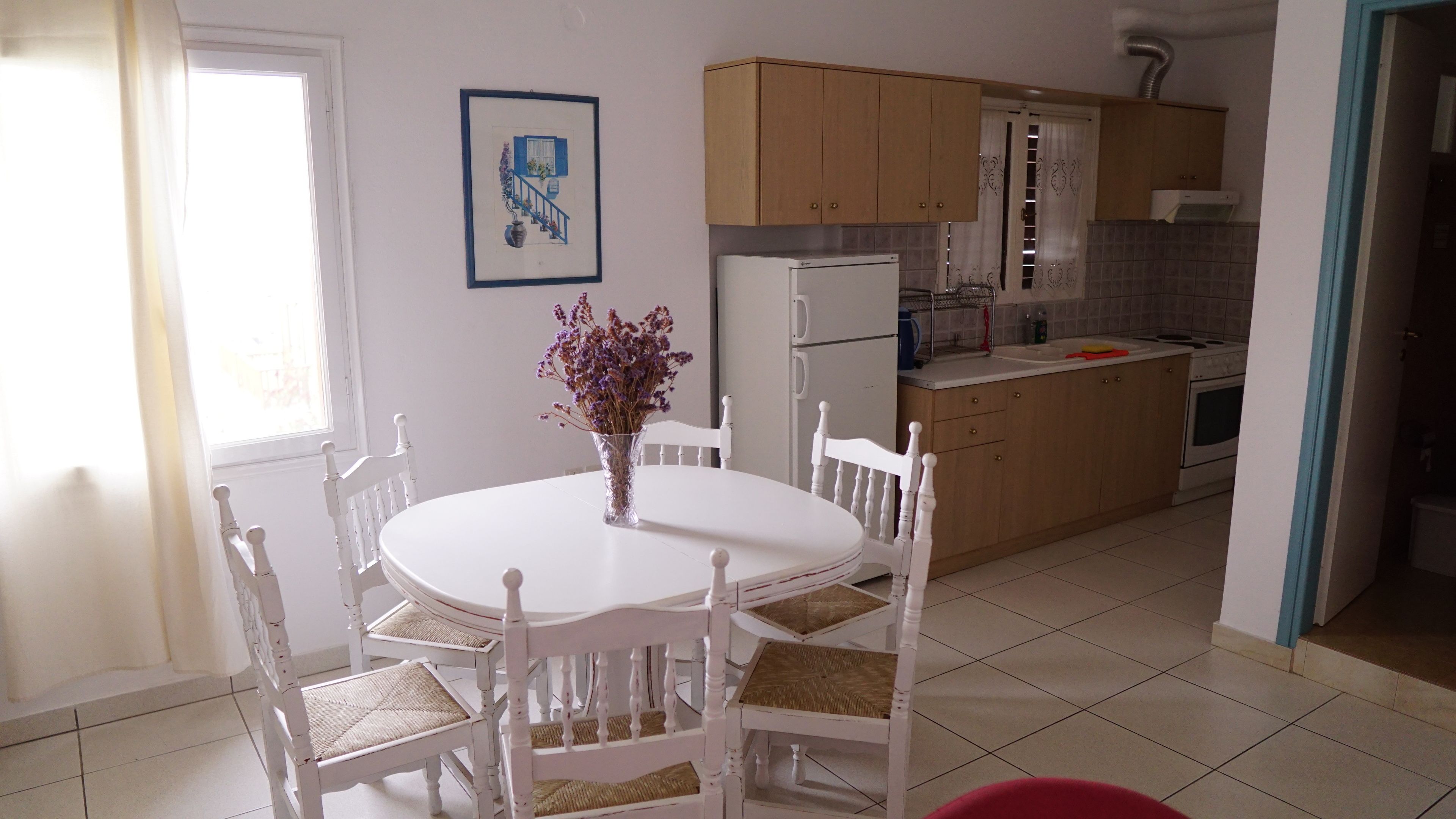 Un comedor con una mesa y sillas blancas, un florero con flores, y una cocina al fondo.