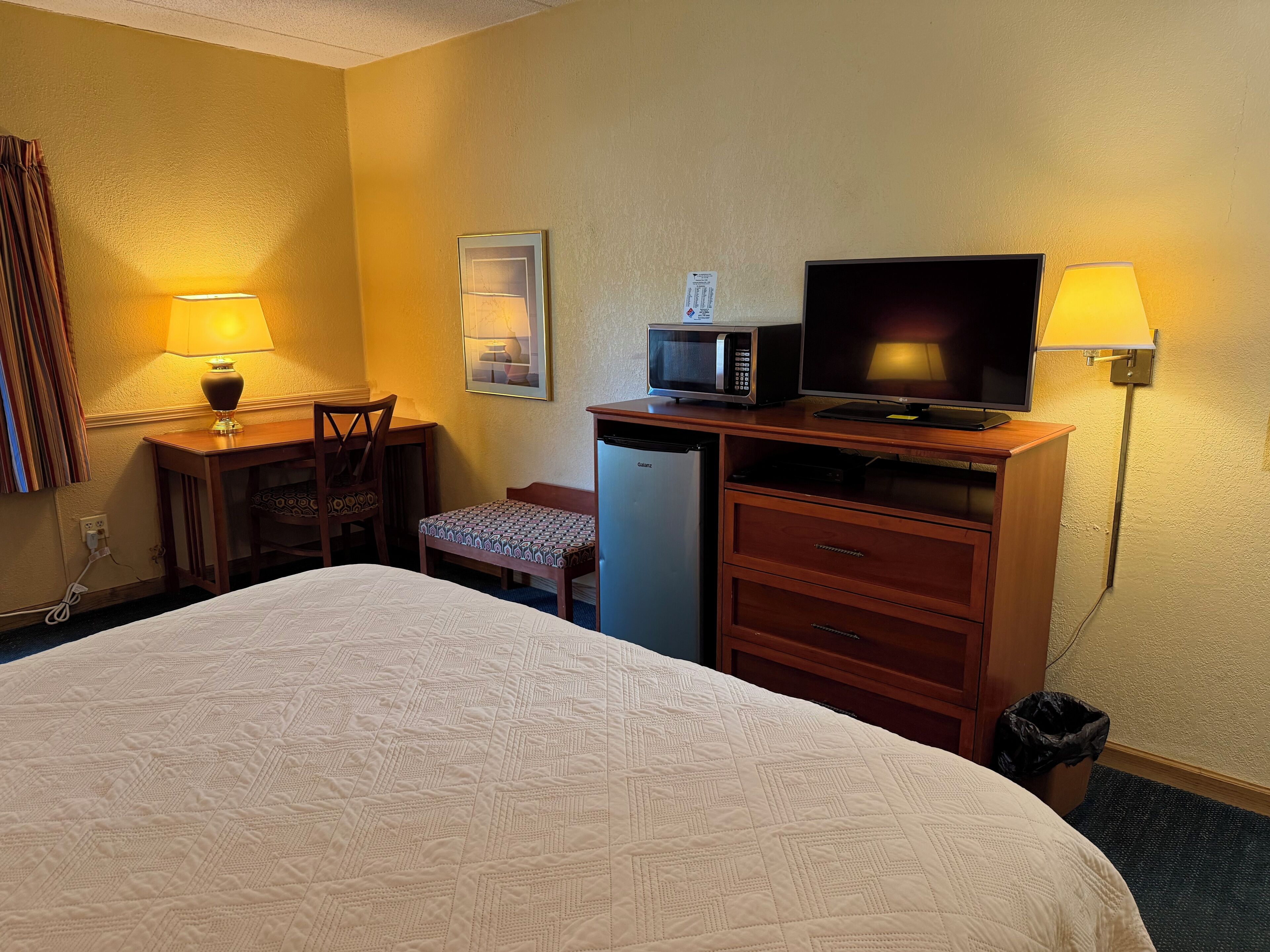 A hotel room with a bed, desk, chair, television, microwave, and a trash bin.
