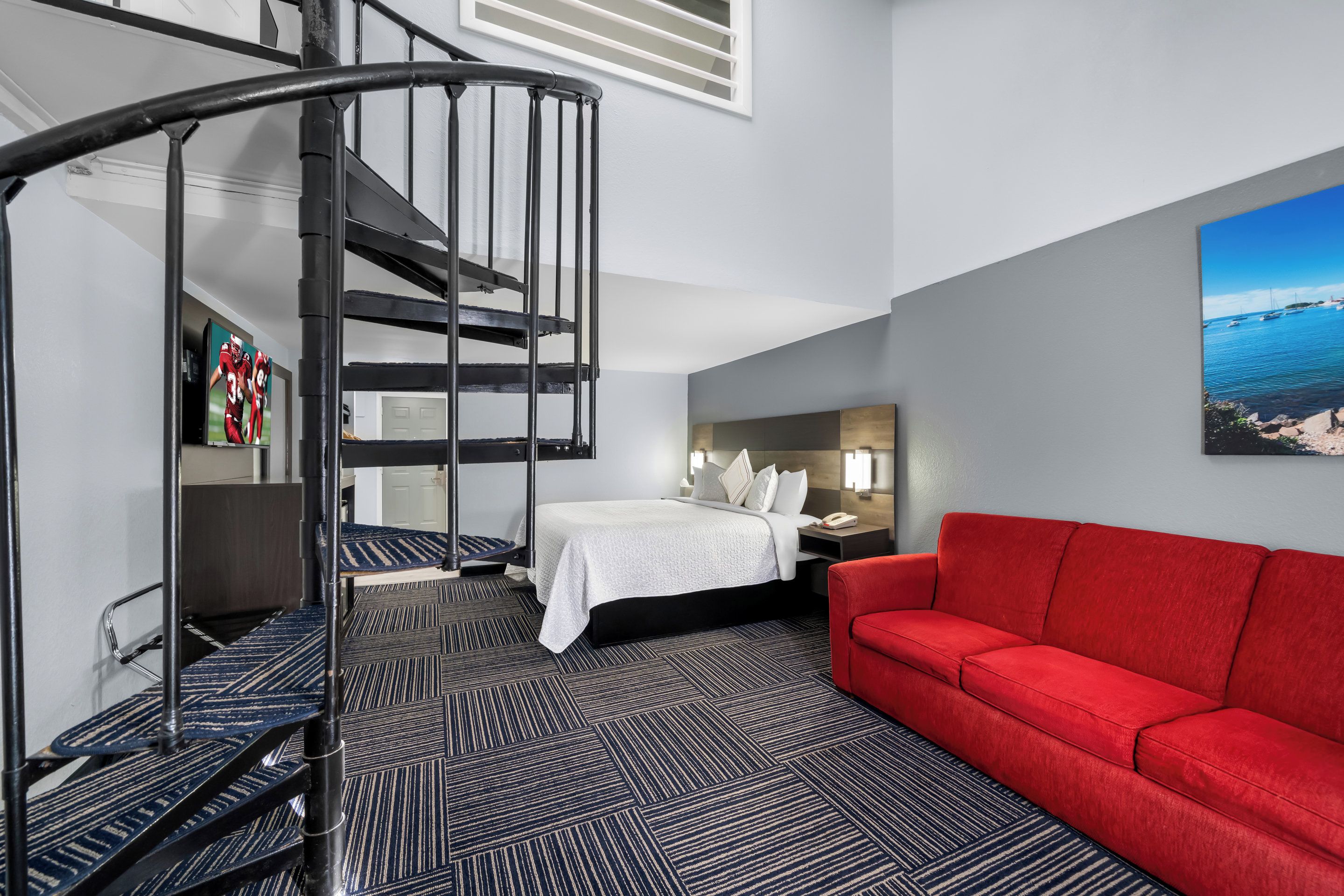 Habitación de hotel con una escalera espiral, una cama, un sofá rojo y un cuadro de una escena costera.