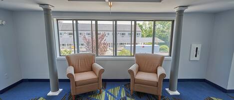 A room with two beige armchairs, a patterned carpet, a large window with a view of buildings, and a ceiling with recessed lighting and a smoke detector.