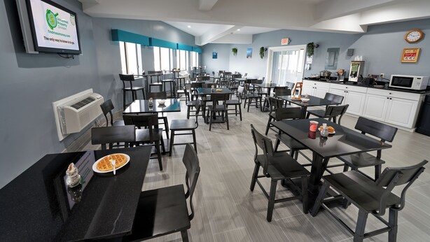 A dining area with multiple tables and chairs, a kitchen area with appliances, and a television displaying a logo. - Admiralty Inn & Suites - a Red Collection Hotel (East Falmouth)