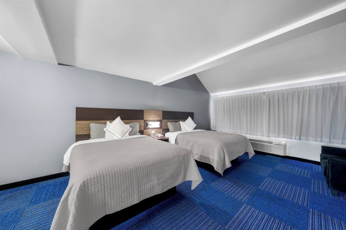 A hotel room with two beds, a blue carpet, a wooden headboard, a nightstand with a lamp, and a window with curtains.