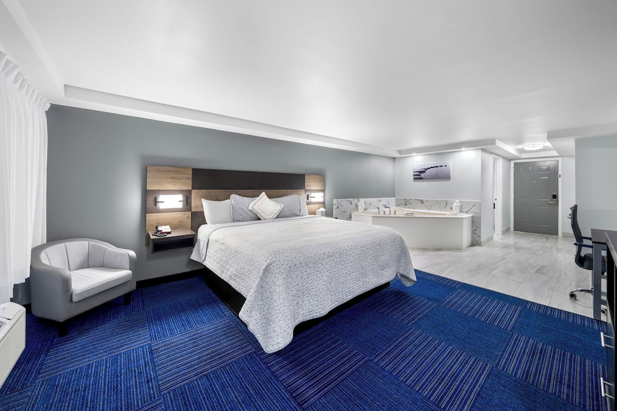 A modern hotel room with a large bed, a sitting area with a chair, a desk, and a bathroom with a bathtub and shower.