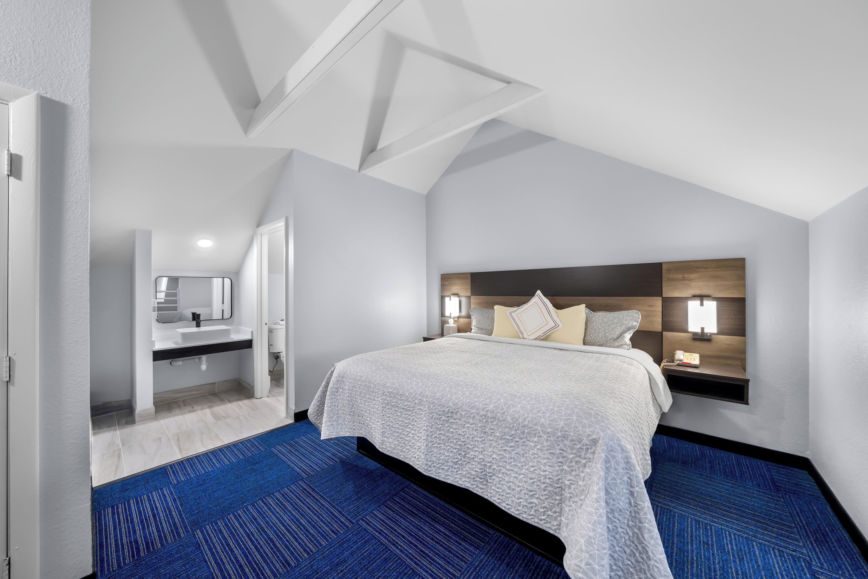 A modern hotel room with a large bed, a nightstand, a wall-mounted lamp, and a bathroom visible through an open door.