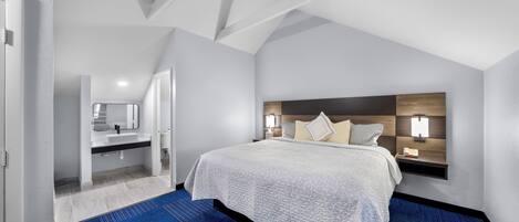 Una habitación de hotel moderna con una cama grande, una mesita de noche, una lámpara de pared y un baño visible a través de una puerta abierta.