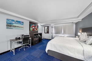 A hotel room with a large bed, a desk with a chair, a TV, a wardrobe, and a window with curtains.