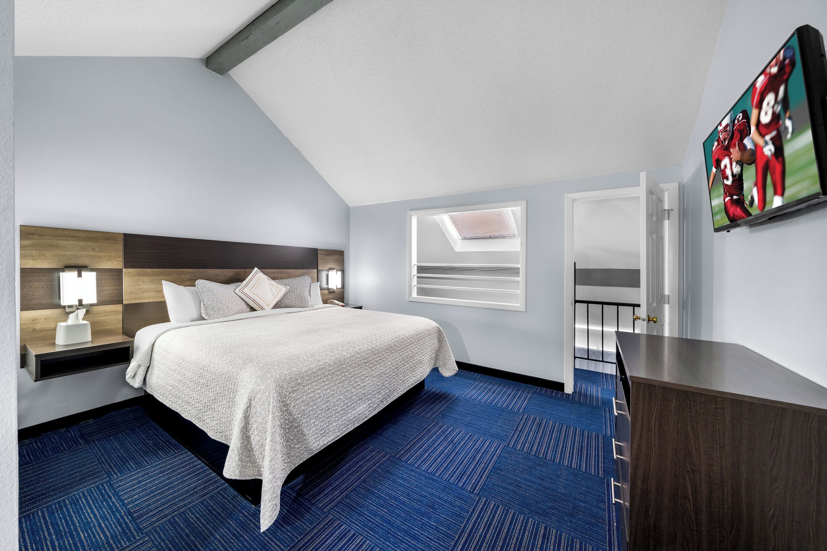 Une chambre d’hôtel moderne avec un grand lit, une table de chevet avec une lampe, un téléviseur fixé au mur et une moquette bleue.