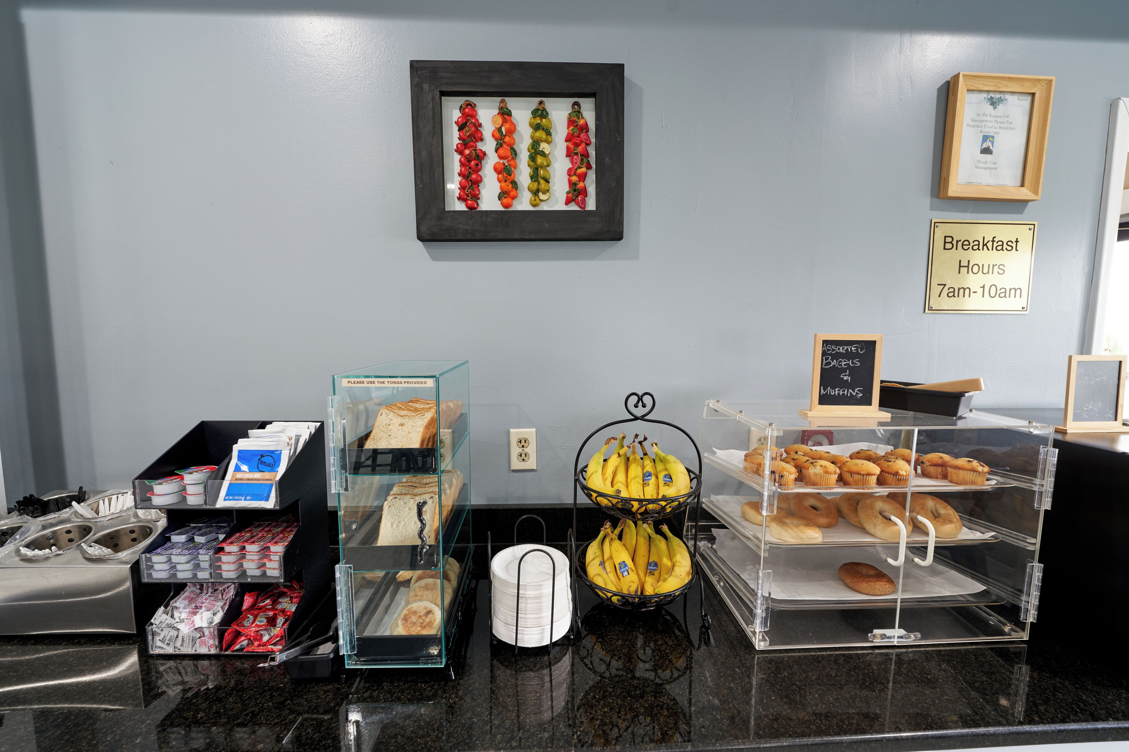 Een ontbijtbuffet met diverse gerechten, een bord met de ontbijttijden en een foto in een lijst aan de muur.