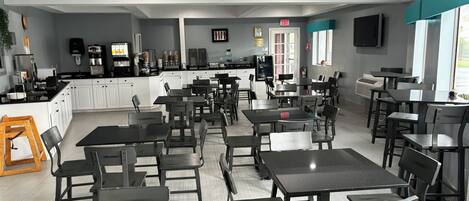 A spacious cafe with multiple tables and chairs, a kitchen area, and a large window providing natural light.