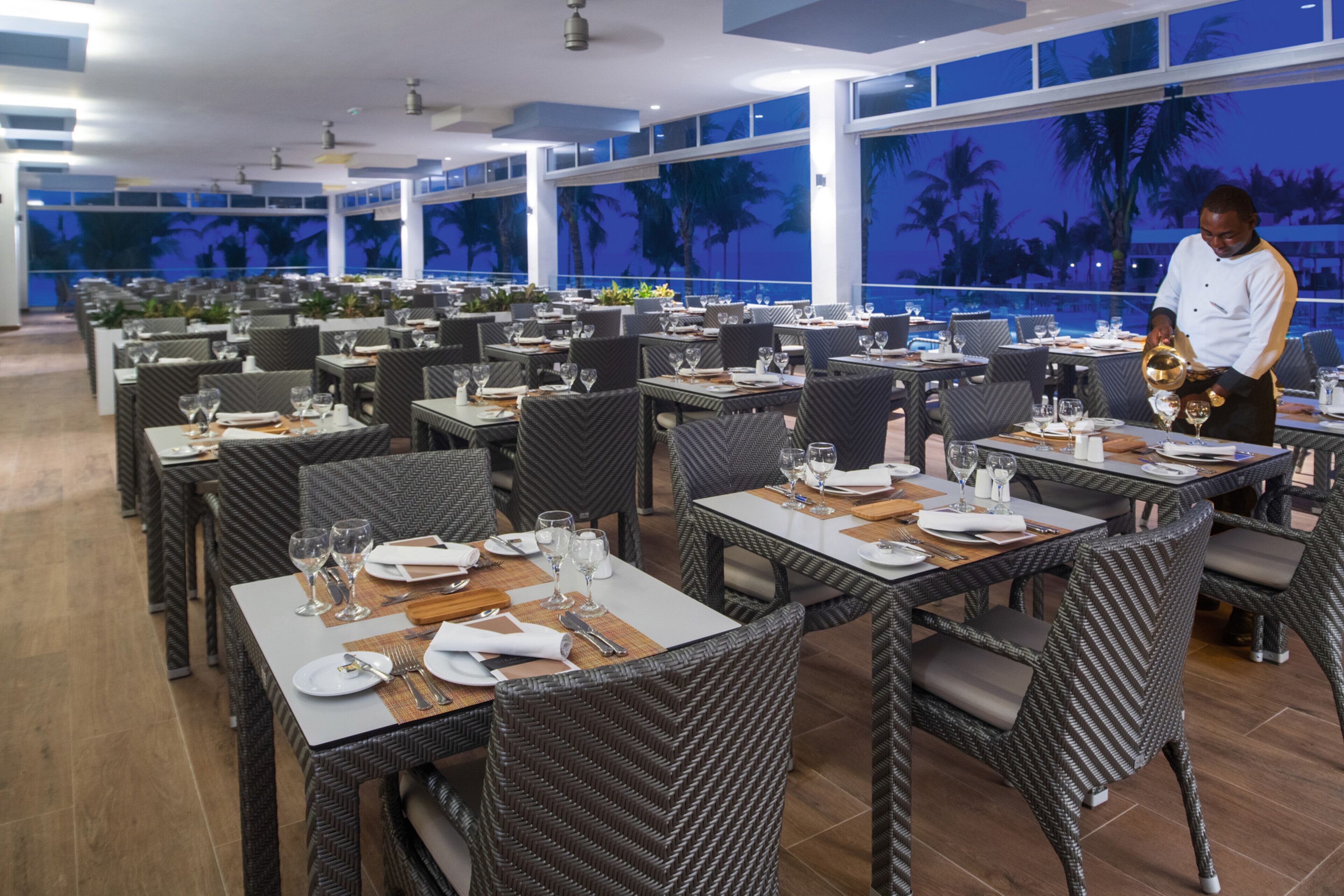A restaurant with multiple tables set for dining, featuring wicker chairs and a view of palm trees through large windows.