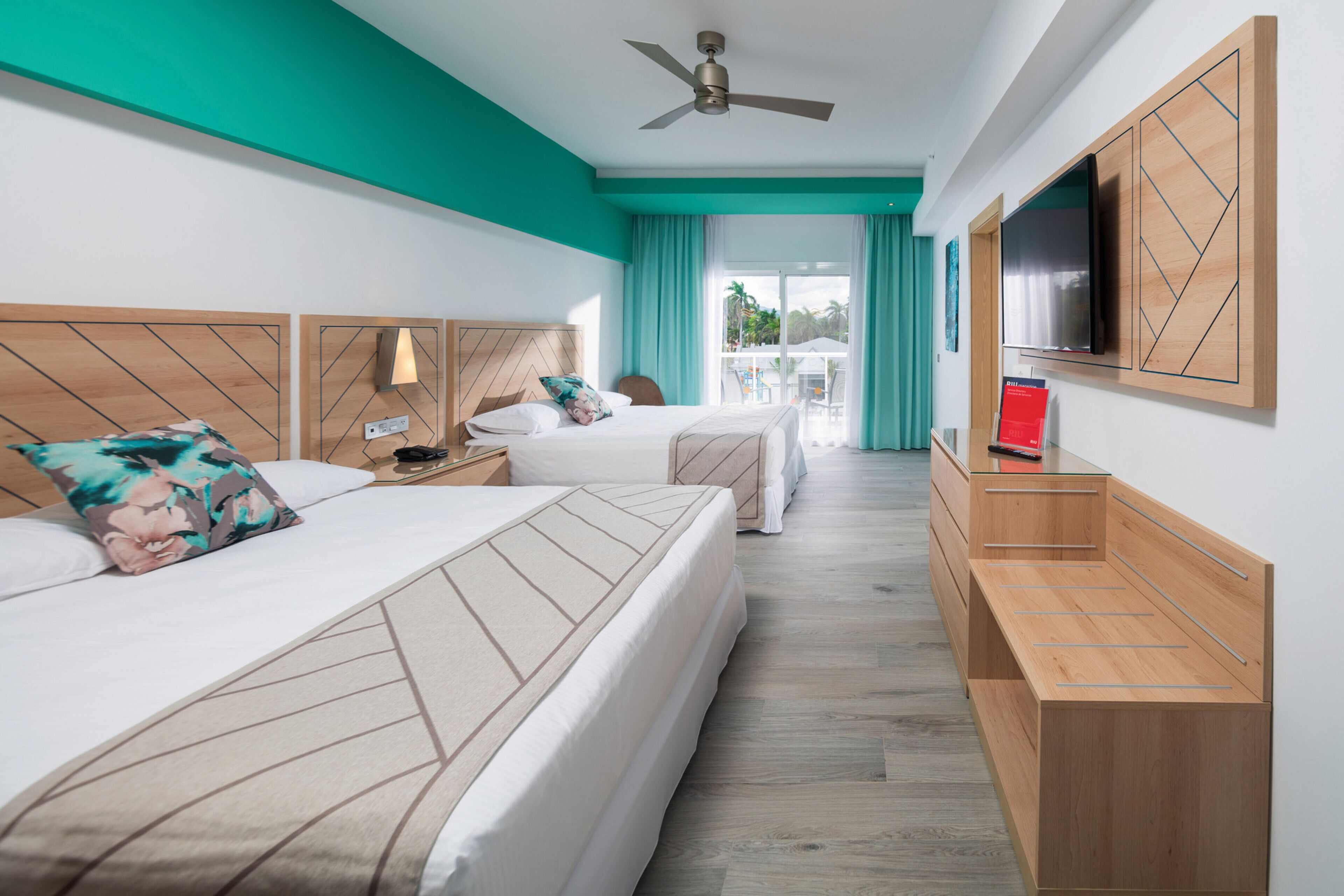 A hotel room with two beds, a ceiling fan, a TV, and a view of the outdoors.
