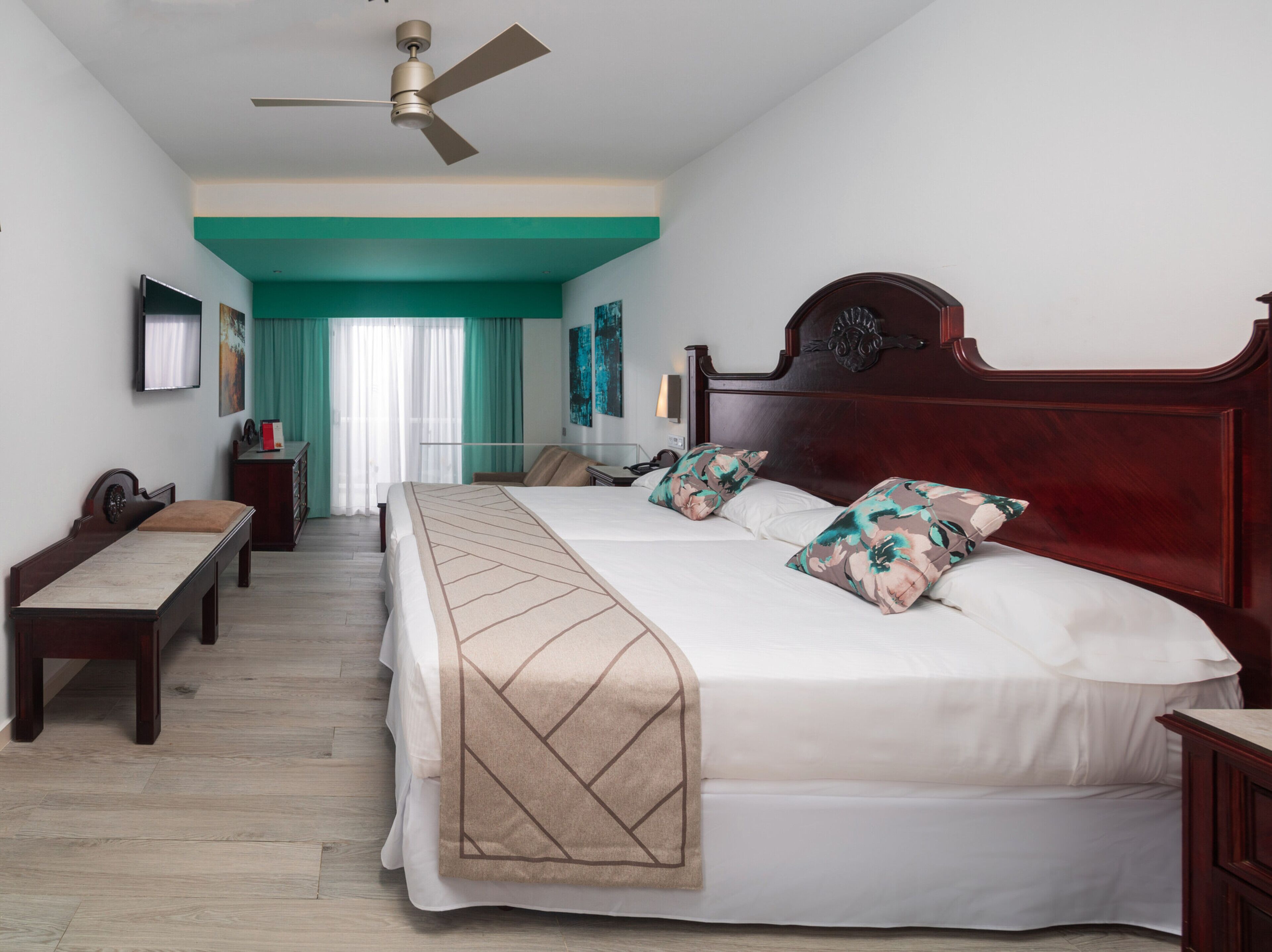A hotel room with a large bed, a desk, a chair, a TV, and a ceiling fan.