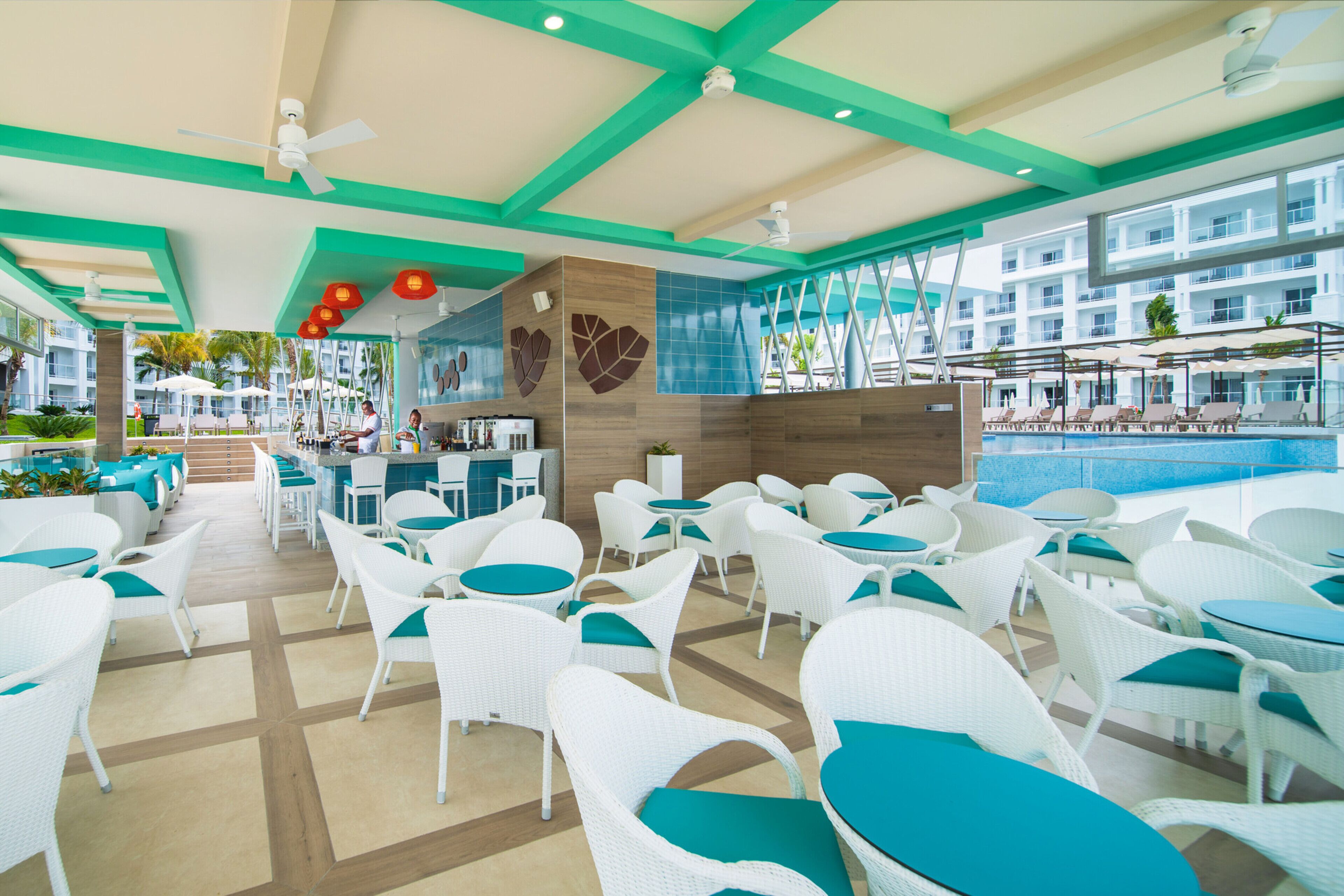 A poolside area with white wicker chairs and tables, a bar area, and a view of the pool and hotel buildings.