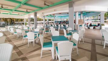 A spacious outdoor dining area with white wicker chairs and green table tops, featuring a checkered floor pattern and a view of the pool area.