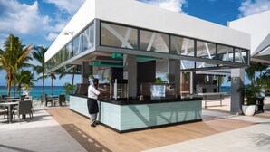 A modern outdoor bar area with a view of the ocean, featuring a counter, seating, and a person working behind the bar. - Hotel Riu Ocho Rios - All Inclusive (Ocho Rios)