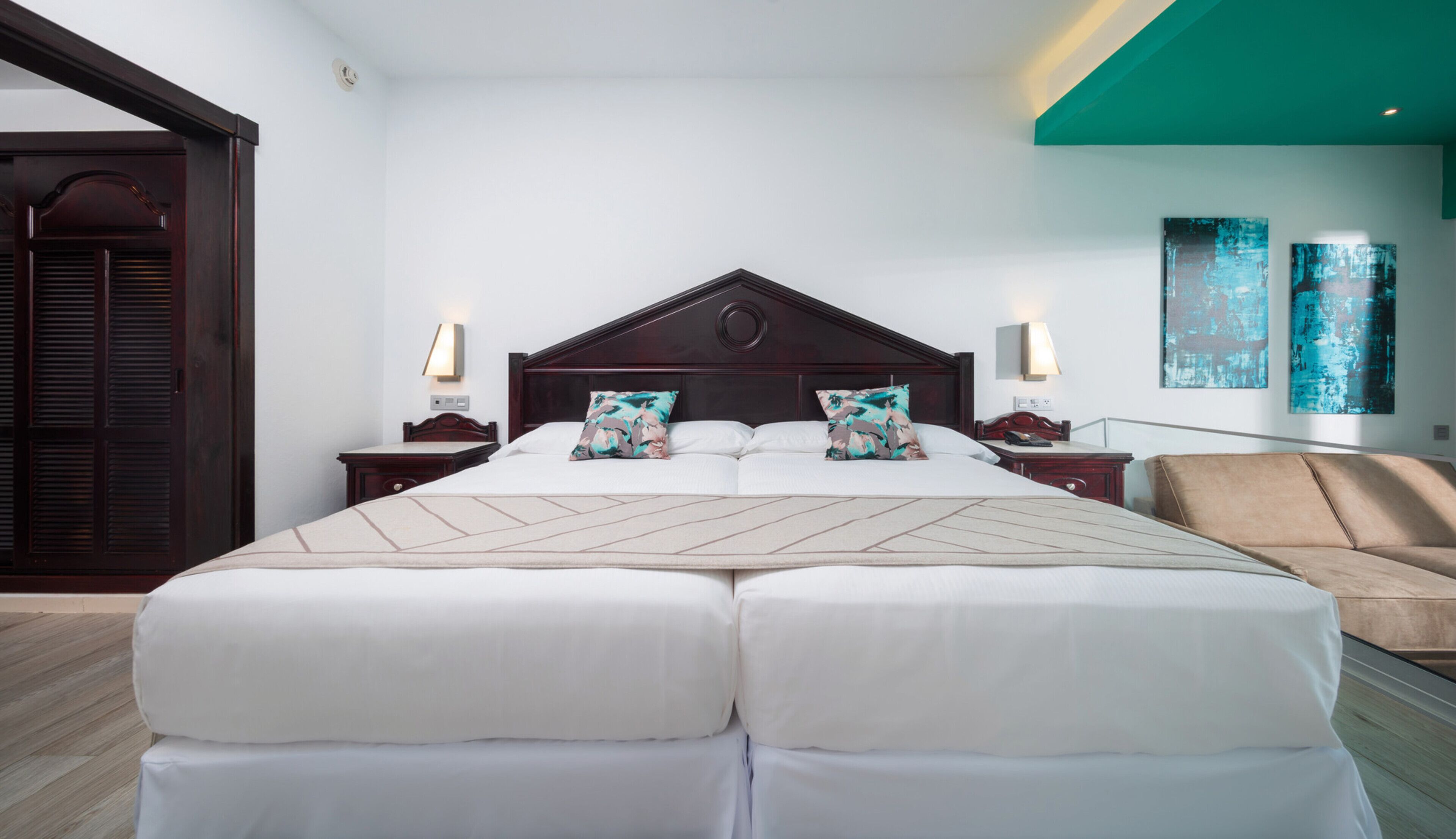 A large bed with a wooden headboard, two bedside tables with lamps, a beige sofa, and abstract wall art.
