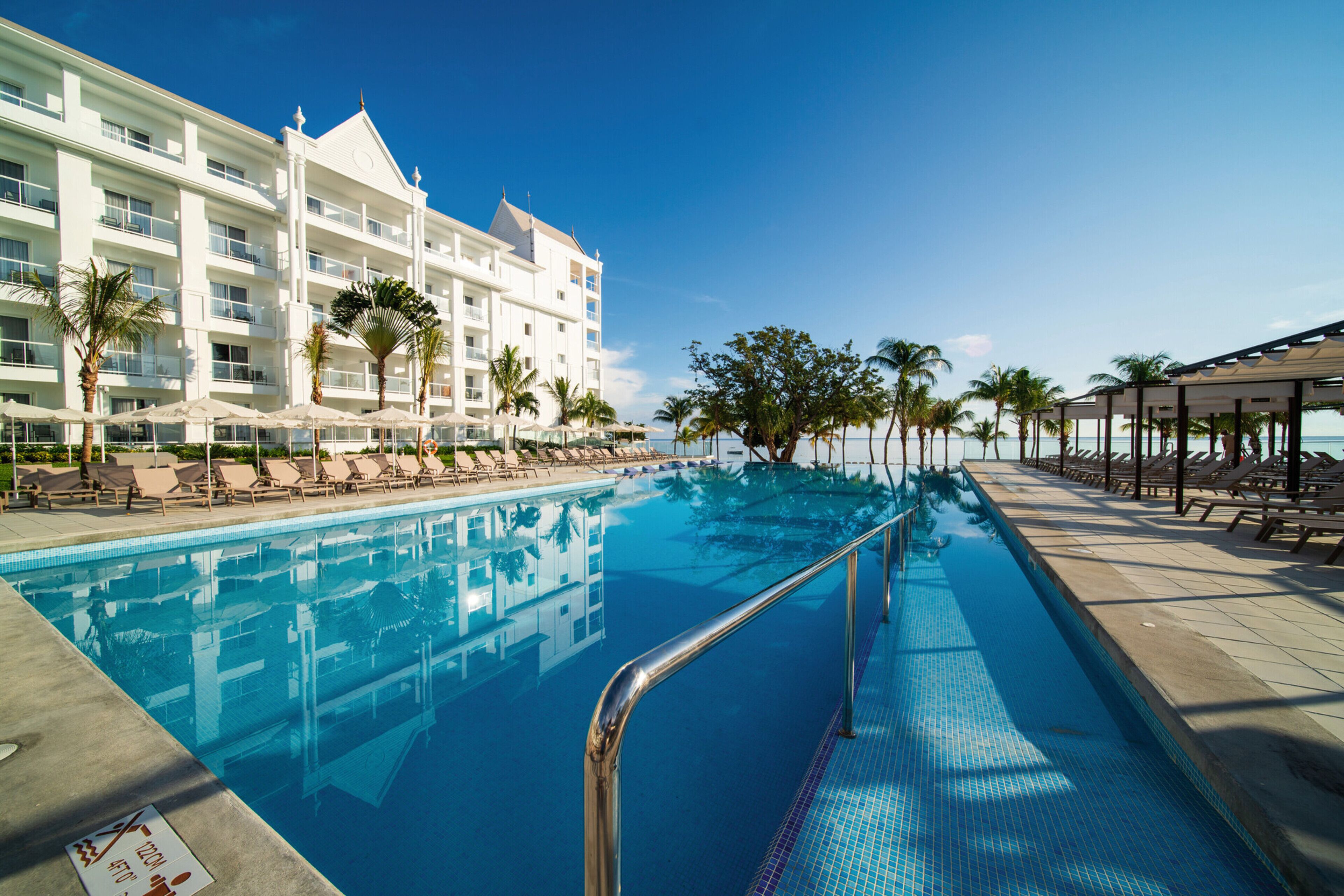 A hotel pool area with a clear blue pool, lounge chairs, palm trees, and a building with multiple balconies.