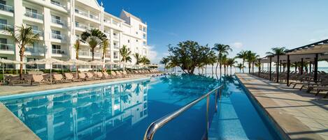 A hotel pool area with a clear blue pool, lounge chairs, palm trees, and a building with multiple balconies.