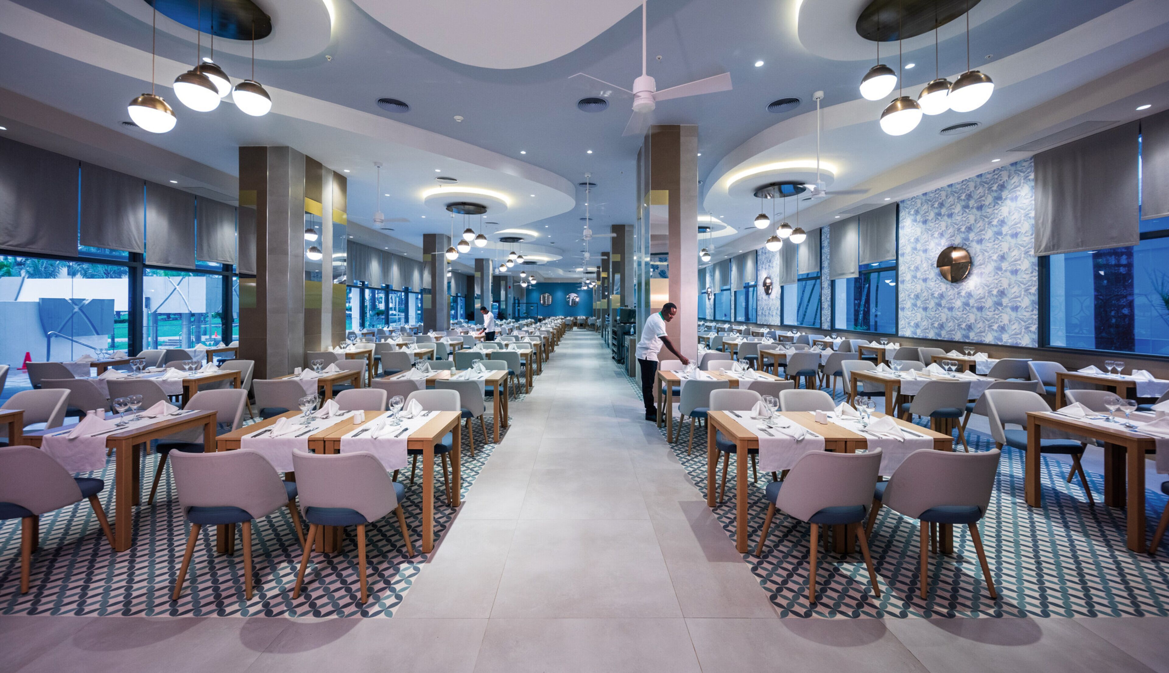 A spacious restaurant with multiple dining tables and chairs, large windows, and modern ceiling lights.