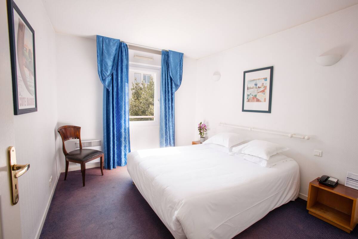 Habitación de hotel con una cama grande, una silla, una mesita de noche y una ventana con cortinas azules.