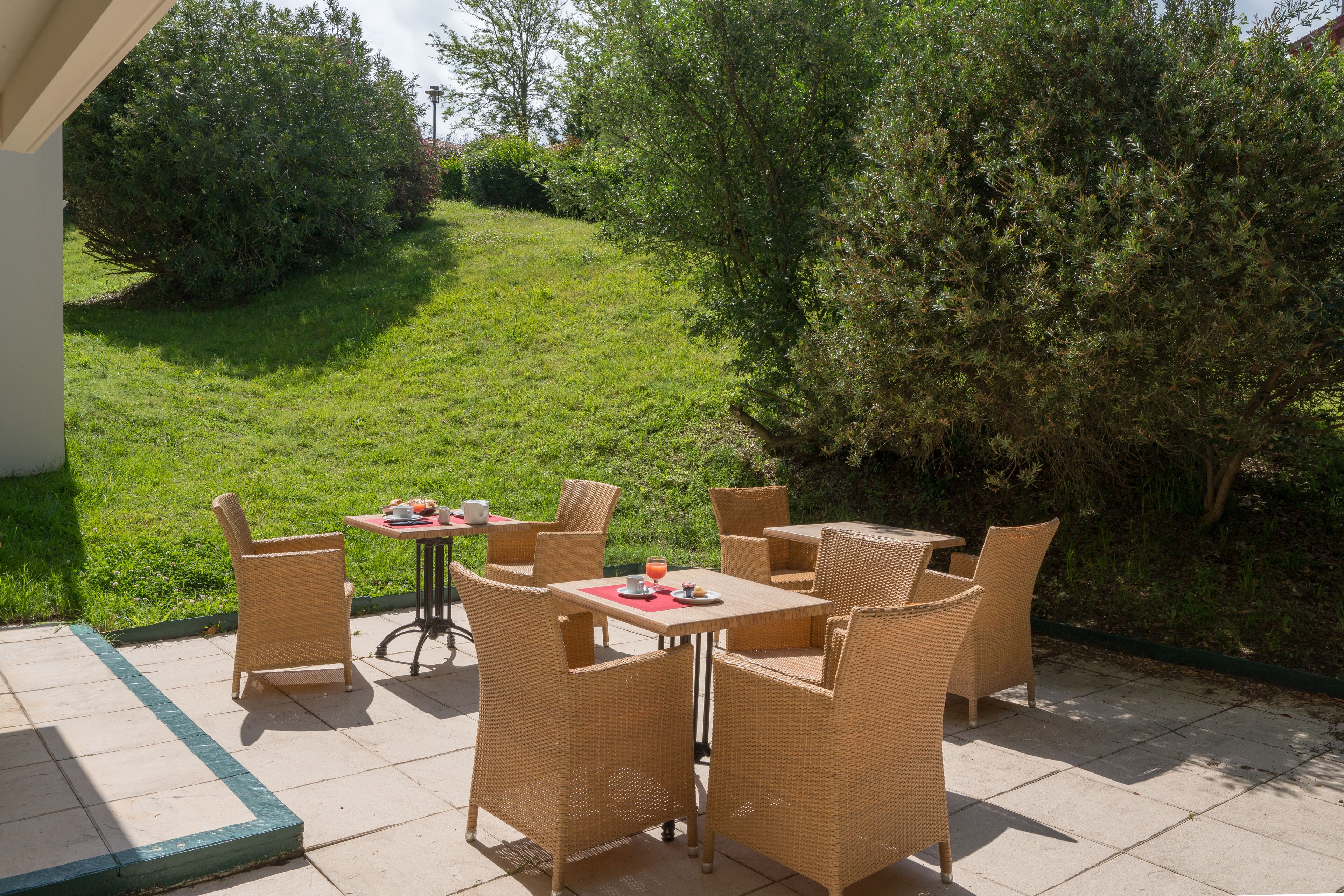 An outdoor patio with wicker furniture and a table set for two, surrounded by greenery.