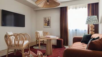 Ein modernes Hotelzimmer mit Flachbildfernseher, Korbstühlen, einem runden Tisch, einer Couch und einem gemusterten Teppich.
