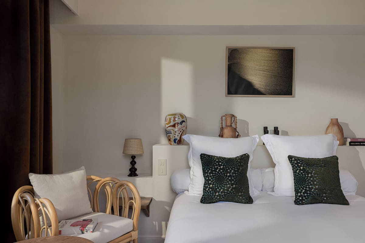 A hotel room with a bed, two pillows, a chair, a nightstand, a lamp, and a wall-mounted artwork.