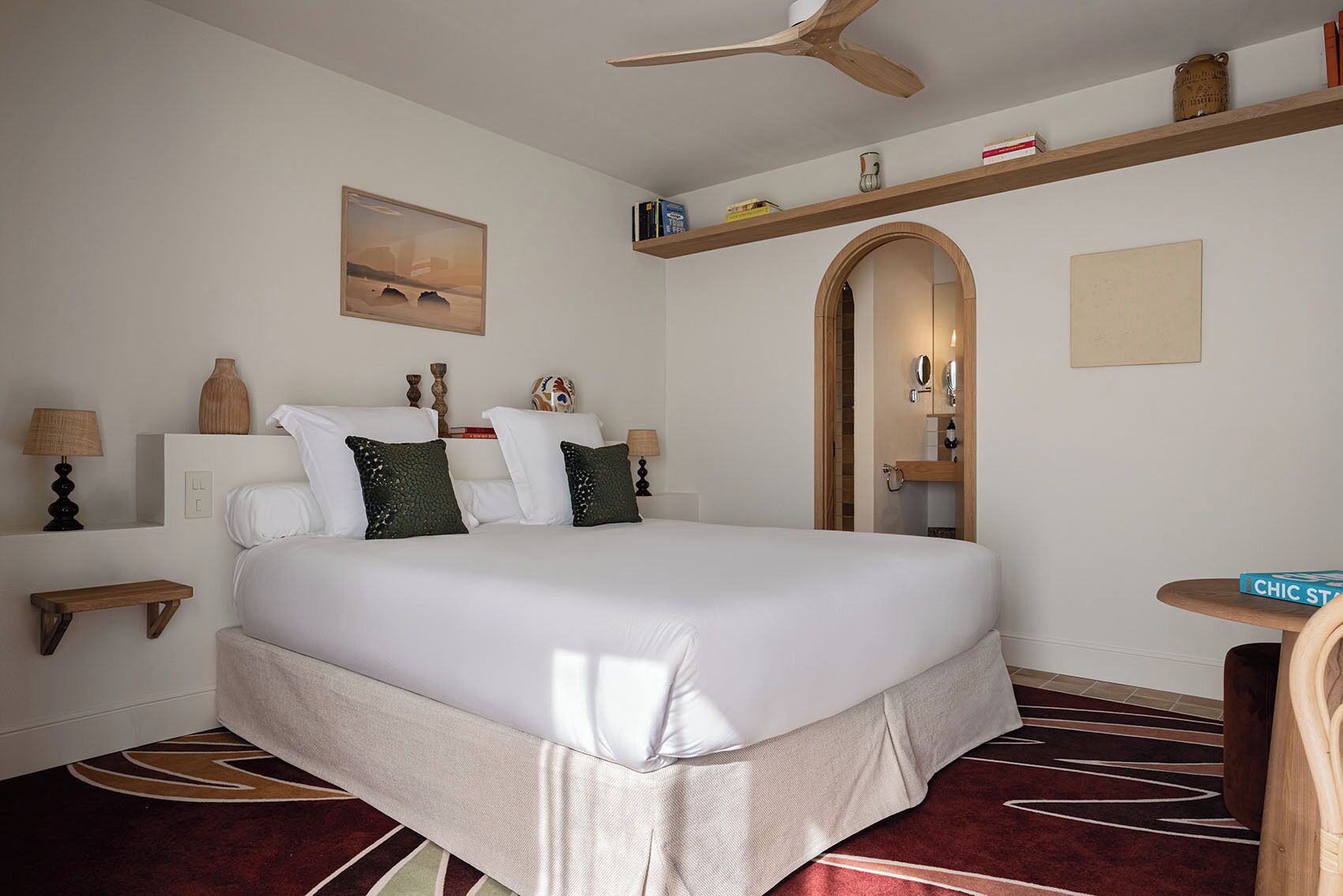A hotel room with a large bed, a ceiling fan, a small table, and a bookshelf.