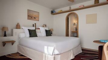 Una habitación de hotel con una cama grande, un ventilador de techo, una mesita pequeña y una estantería.