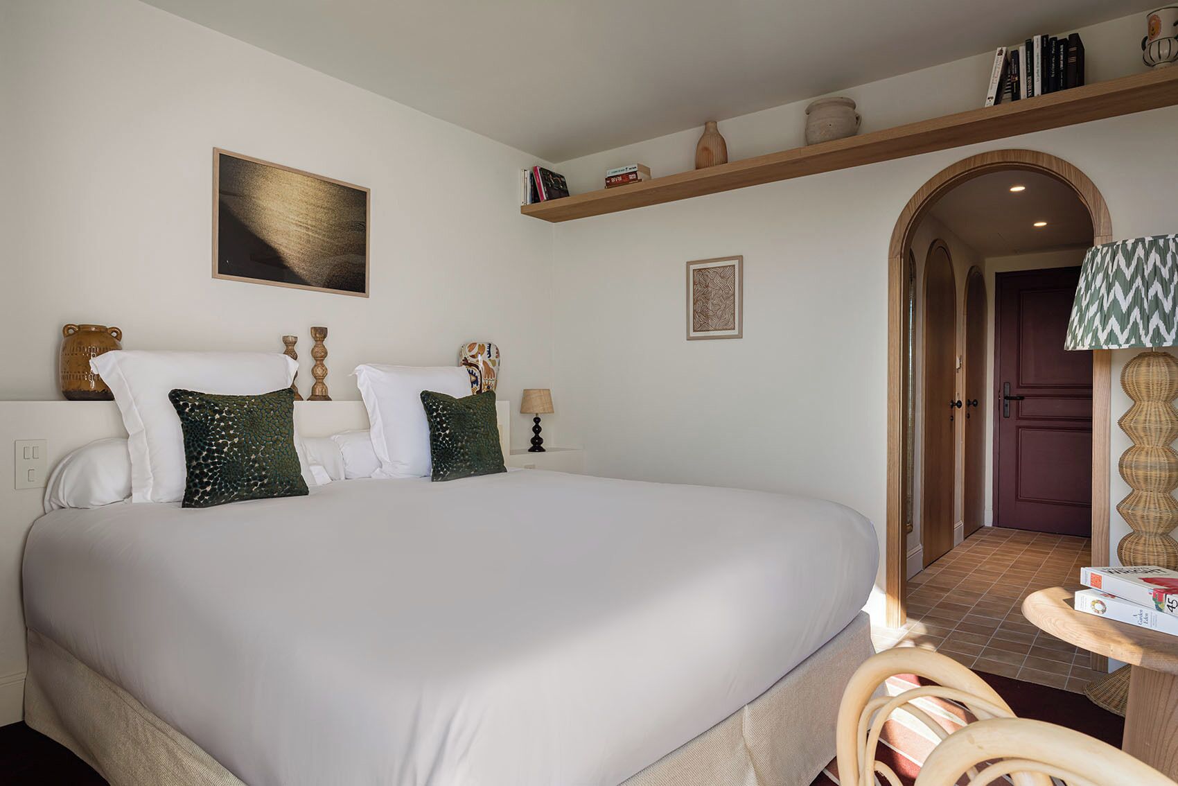 A neatly arranged bedroom with a large bed, a bedside table, a lamp, and a shelf with books and decorative items.