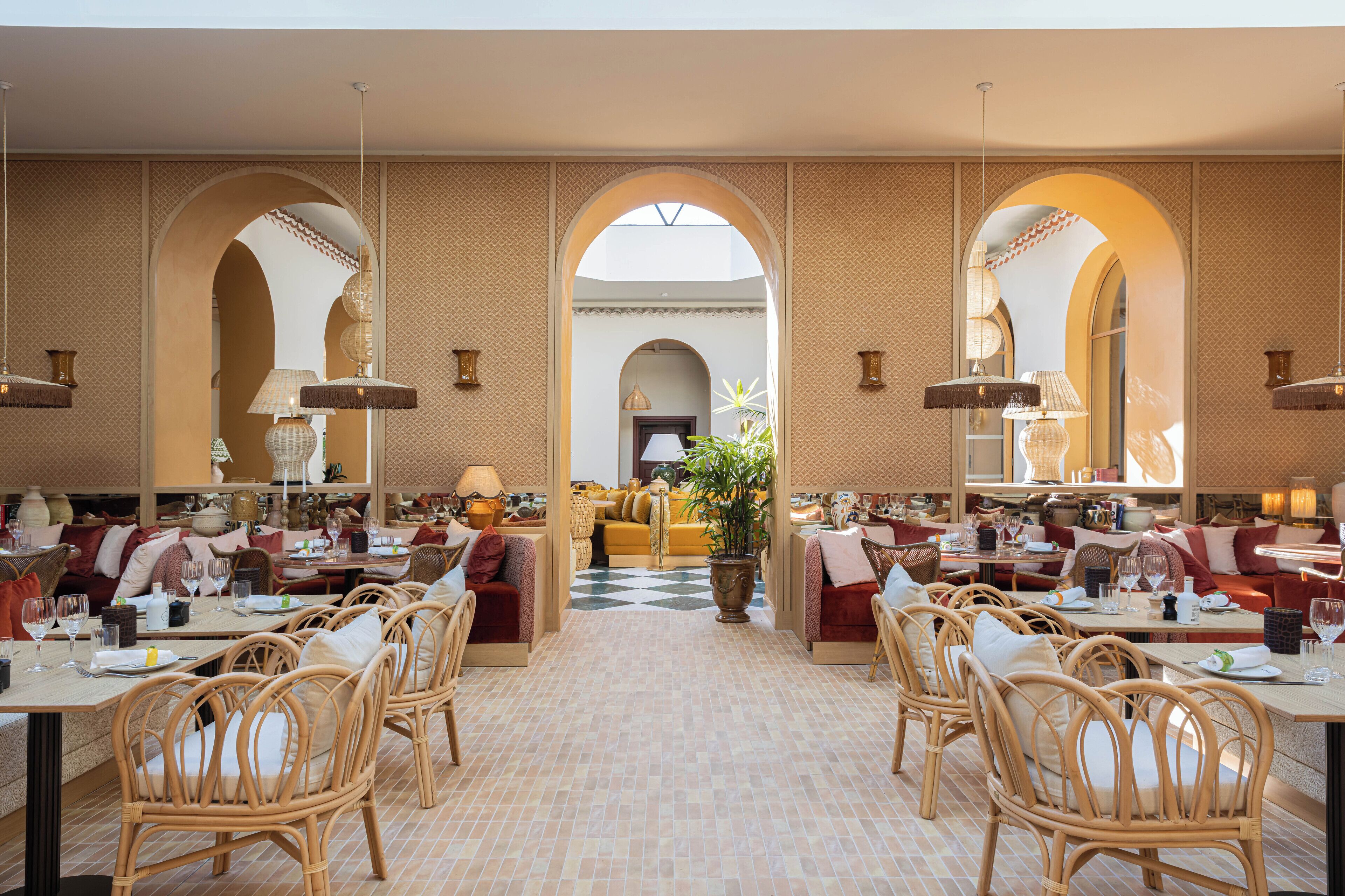 Une salle à manger spacieuse avec de hauts plafonds, des portes en arc de cercle et des meubles en osier. Les tables sont dressées avec des assiettes, des verres et des couverts.