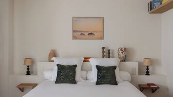A hotel room with a large bed, two decorative pillows, a painting of a landscape, and a bookshelf with books.