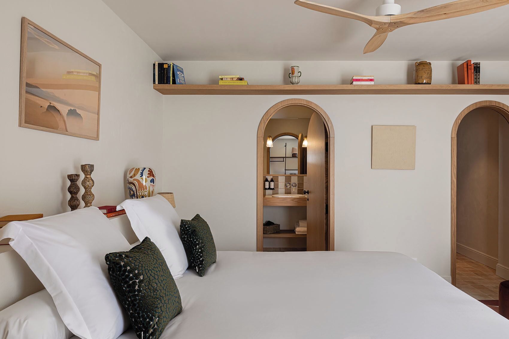 Un dormitorio con cama, ventilador de techo, estantería y un arco que da a un baño.