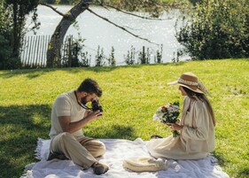 A couple sitting on a blanket by a lake, with a camera and flowers.