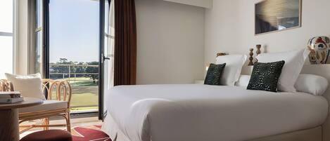 A hotel room with a large bed, a desk with a chair, a view of a golf course through an open window, and a painting on the wall.