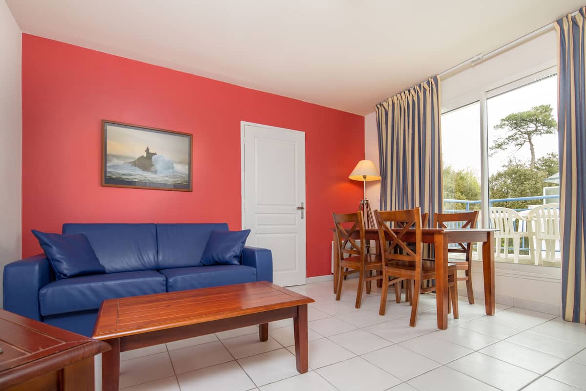 Una habitación con un sofá azul, una mesa de centro de madera, una mesa de comedor con sillas, una lámpara y un cuadro en la pared.