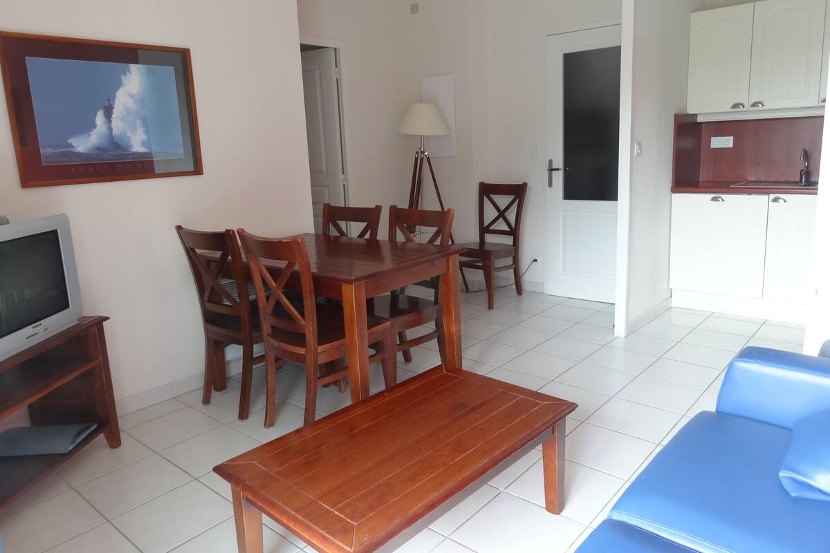 Un comedor con mesa y sillas de madera, un televisor sobre un mueble, una lámpara y una zona de cocina con armarios.