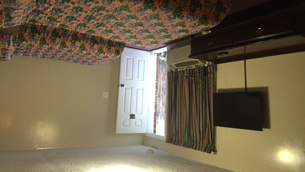 A room with a patterned ceiling, a white door, a window with curtains, and a mounted television. - Blue Falls Motel (Tonawanda)