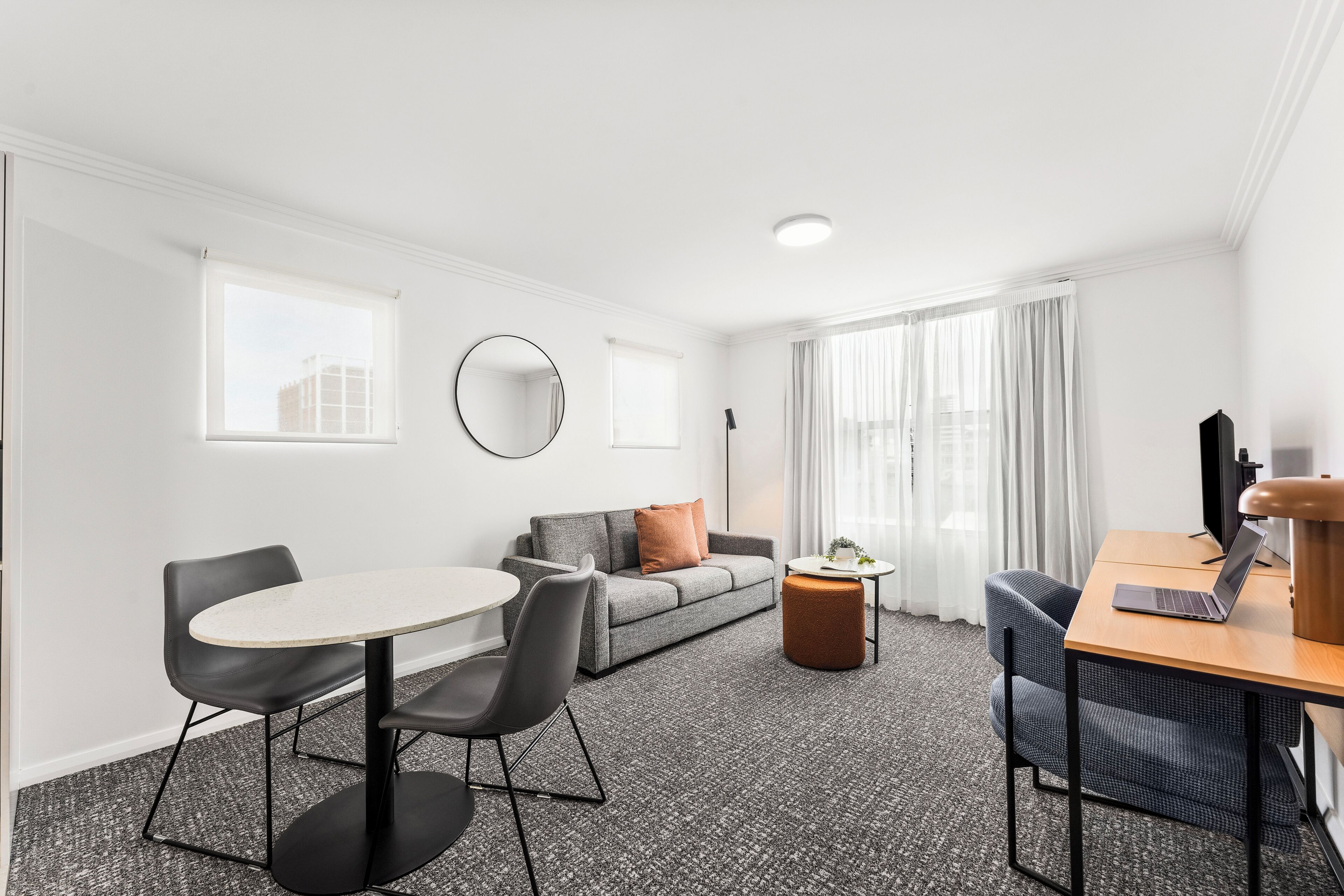 Une chambre d’hôtel moderne avec une table de salle à manger ronde, deux chaises, un canapé, un petit pouf, un bureau avec un ordinateur portable et un grand miroir.