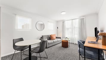 Una habitación de hotel moderna con una mesa redonda para comer, dos sillas, un sofá, un pequeño otoman, un escritorio con una computadora portátil y un espejo grande.