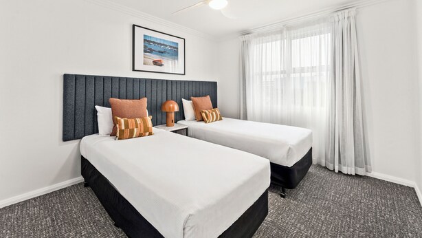 A hotel room with two beds, a ceiling fan, a framed picture on the wall, and a window with sheer curtains.