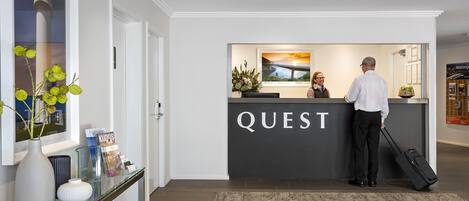 A reception desk with a "QUEST" sign, a vase with yellow flowers, and a framed picture of a sunset over a bridge.