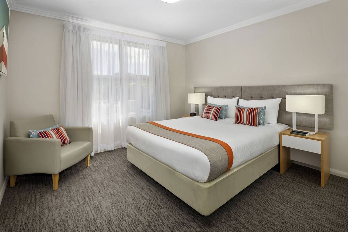 Une chambre d’hôtel avec un grand lit, une chaise, une table de chevet avec une lampe et une fenêtre avec des rideaux légers.