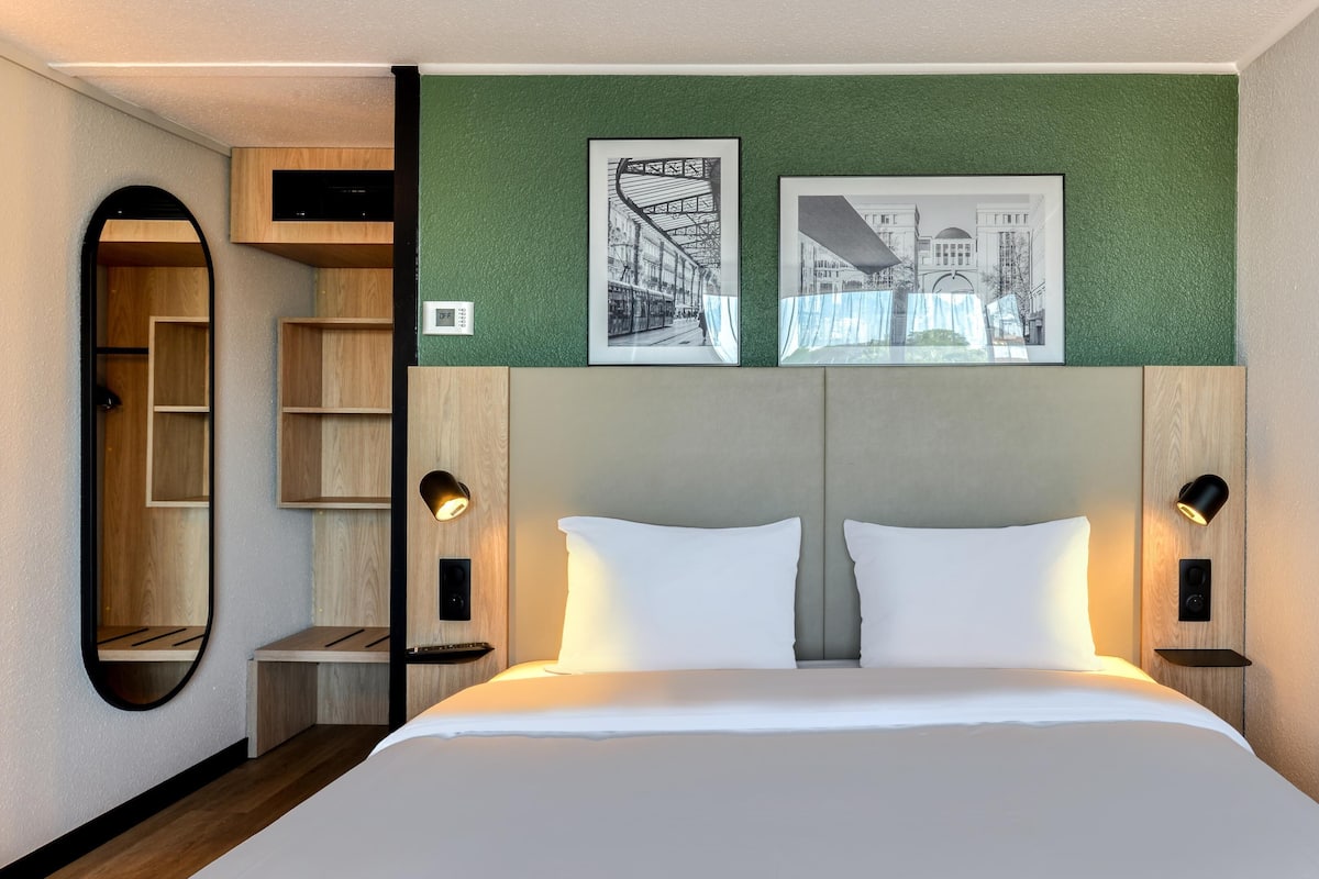 Une chambre d’hôtel comprenant un lit, deux tables de chevet avec des lampes, un grand miroir, une étagère en bois et deux tableaux encadrés accrochés au mur.