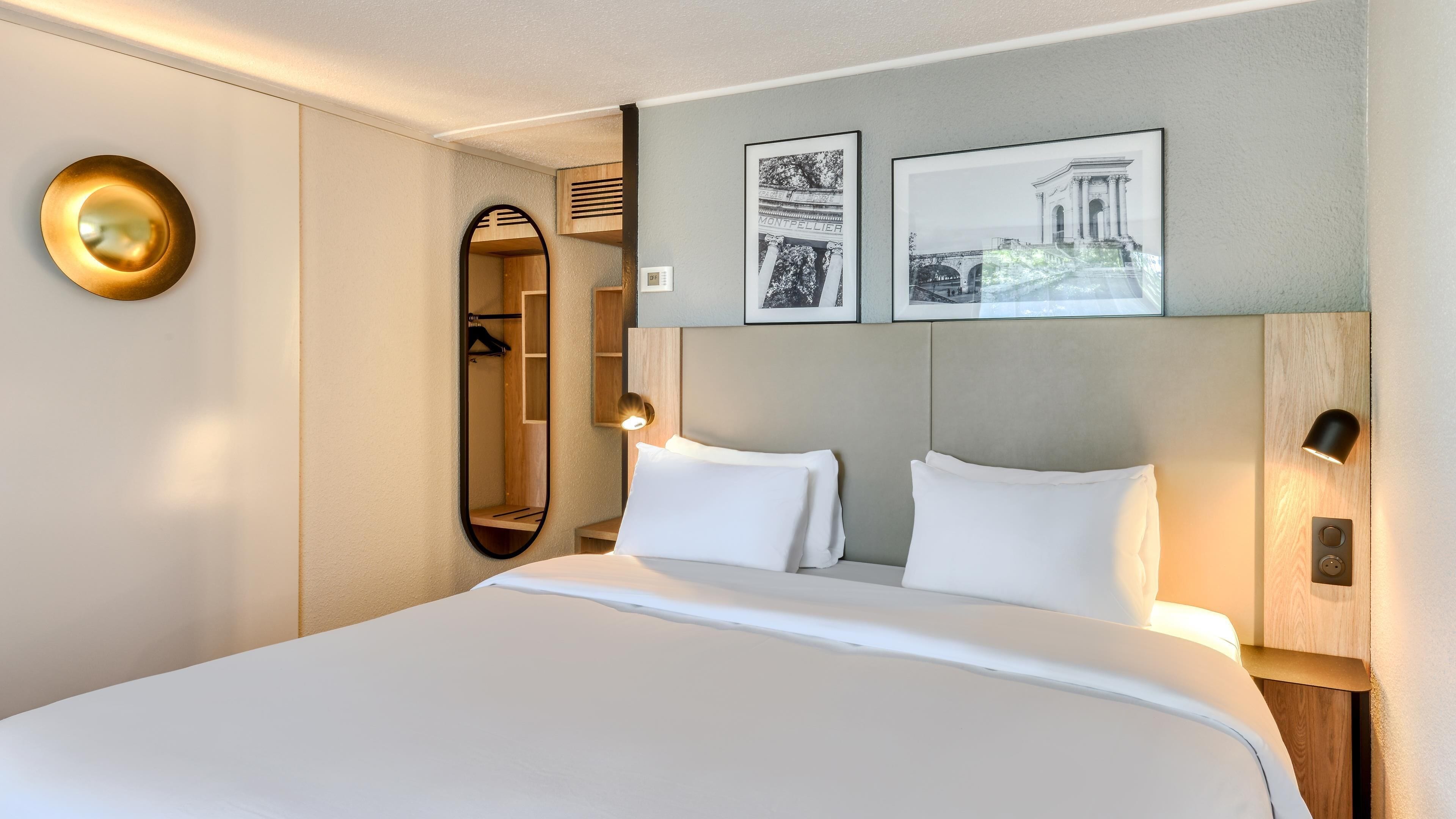 A hotel room with a large bed, a mirror, two framed pictures on the wall, and a wooden headboard.