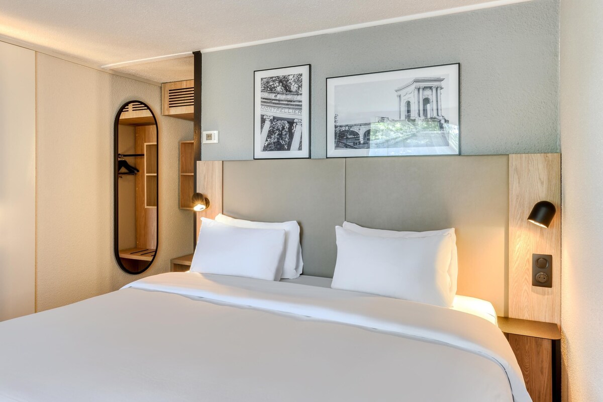 Une chambre d’hôtel avec un grand lit, un miroir, deux tableaux encadrés au mur et une tête de lit en bois.