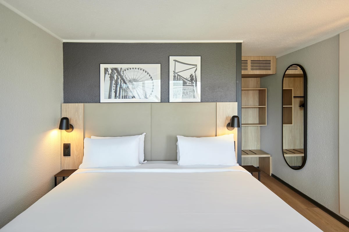 Une chambre d’hôtel avec un grand lit, deux lampes fixées au mur, un miroir et deux tableaux encadrés.