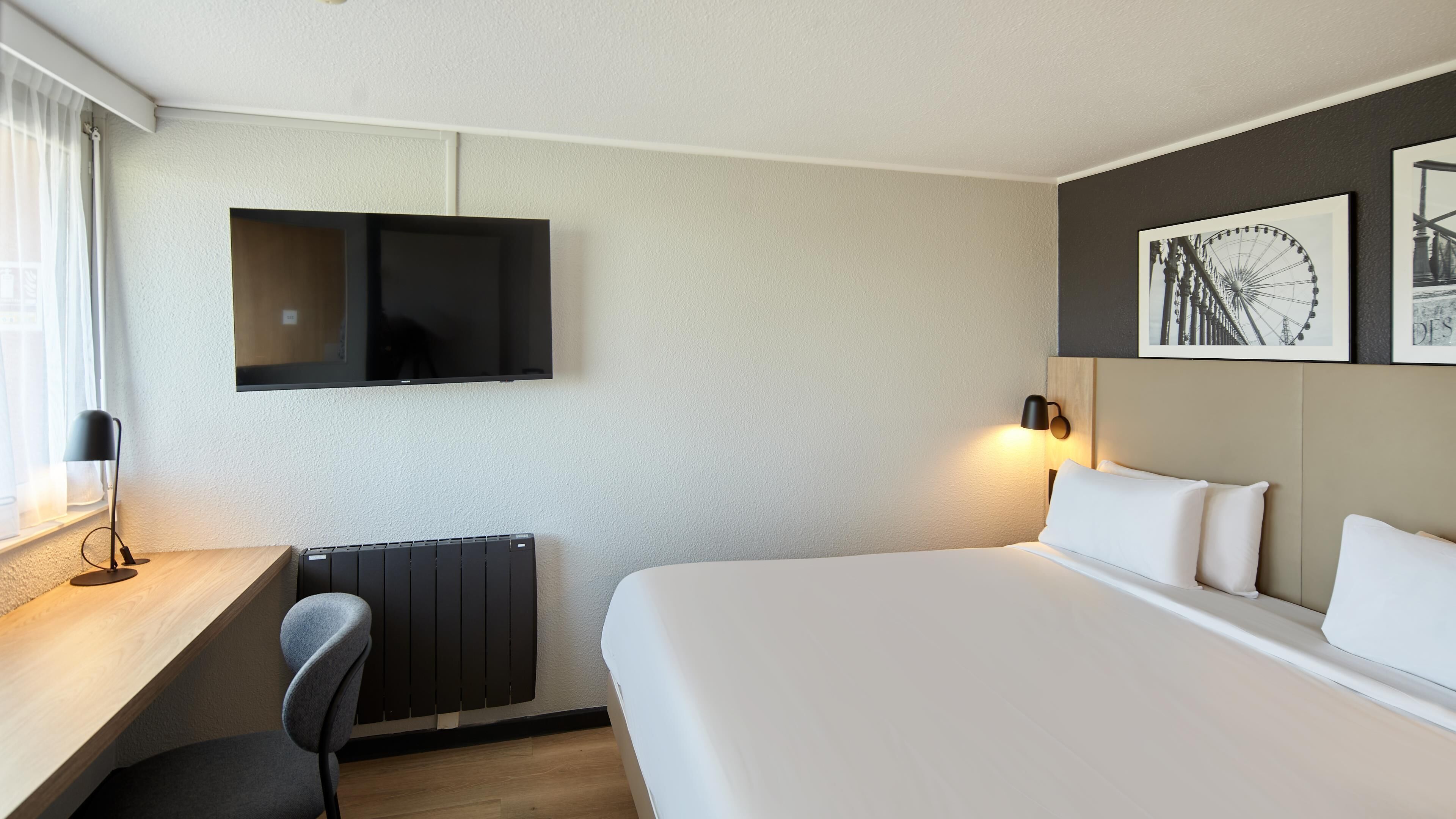 A hotel room with a bed, a desk, a chair, a TV mounted on the wall, and a wall-mounted lamp.
