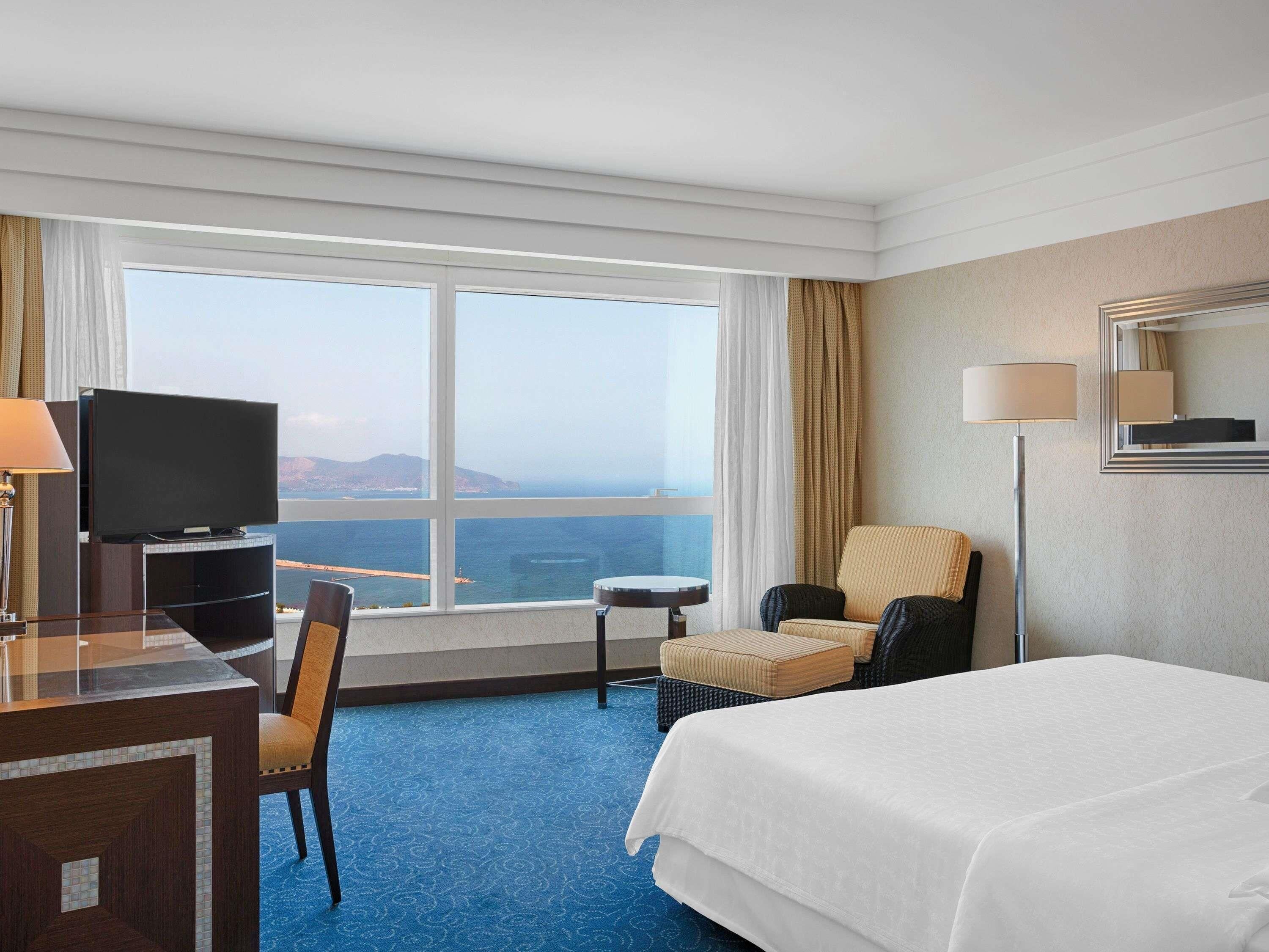 Une chambre d’hôtel avec un lit, un bureau avec une télévision, une chaise, une petite table et une vue sur la mer.